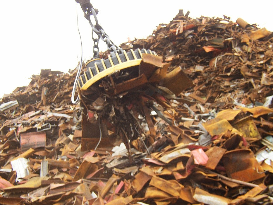 A scrap handling magnet with a yellow and black structure, attached to a crane, surrounded by piles of rusty metal scrap.